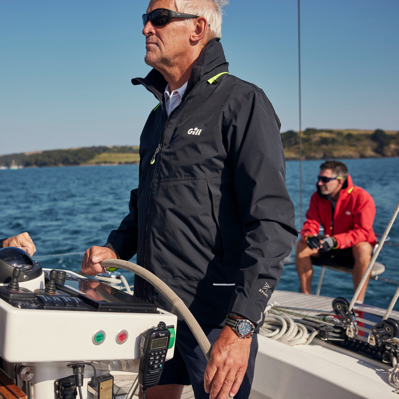 Man steering a sailboat with another person in the background on a clear day.